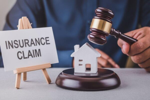 Ręka trzymająca młotek sędziowski obok modelu domu i tabliczki z napisem "insurance claim" symbolizuje zadośćuczynienie za szkody majątkowe.